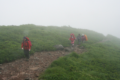 霧にまかれ風が強い山頂直下を歩いている写真