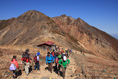 峰の茶屋跡避難小屋と朝日岳を背に歩いている写真