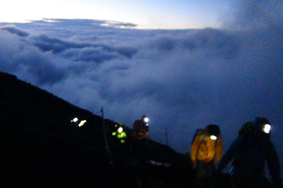 白んできた空の下、雲海を下に見てヘッドランプを付けて登っている写真