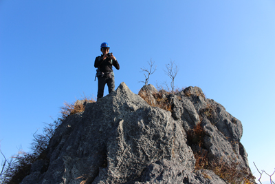 青空をバックに岩場の上に立つMさんの写真