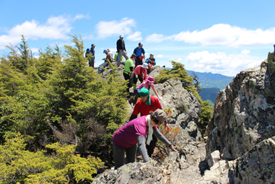 片側がすっぱり切れ落ちた岩場を歩いて山頂に向かうメンバーの写真