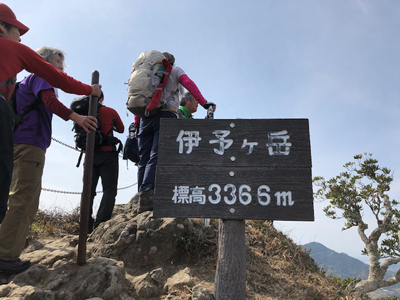 伊予ヶ岳山頂標識の裏で展望を見るメンバーの写真