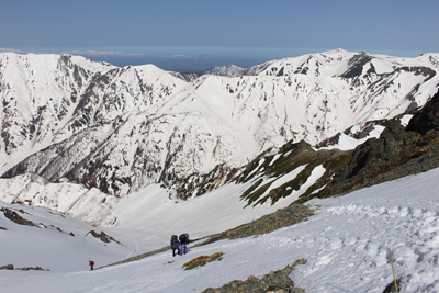双六岳や黒部五郎岳の見える好展望の雪の斜面を登っている写真